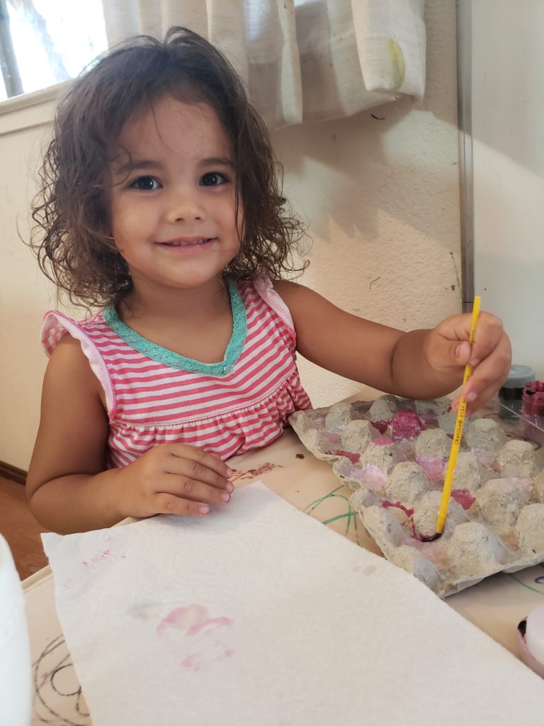 Ana Lia at 3 years old staring at the camera while painting an upside down egg carton