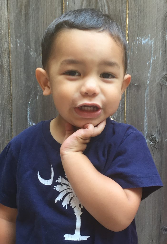 Eliam at 2 years old with a blue shirt and his hand holding his face with a smile