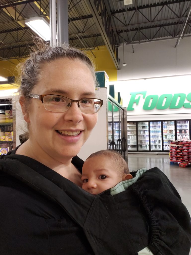 Tromila is in the grocery store with newborn Fin in a chest carrier with both looking into the camera