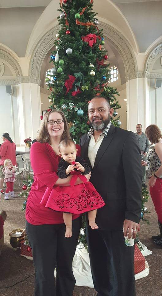 Tromila holding Ana Lia who is 9 months old standing beside Afa in front of a large Christmas tree