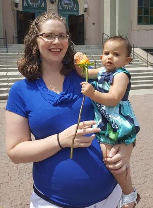 Tromila in a bright blue shirt with a baby bump holding Ana Lia who is holding a yellow rose